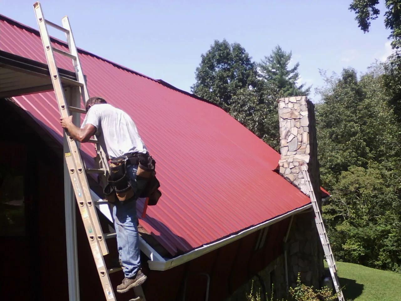 Roofer on a ladder beside a red metal roof and stone chimney — Soffit & Fascia Repair by Roofing Brothers in San Gregorio